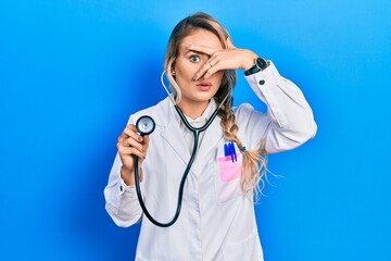 Beautiful young blonde doctor woman holding stethoscope peeking in shock covering face and eyes with hand, looking through fingers with embarrassed expression.