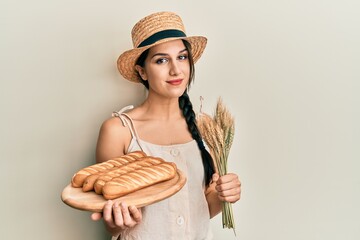 Young hispanic woman holding homemade bread and spike wheat relaxed with serious expression on...