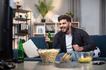 Fototapeta na wymiar Excited indian man with chips and beer sitting on cozy sofa and watching sport TV. Young bearded guy supporting favorite football team during world cup.