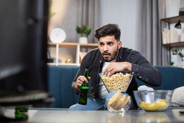 Fototapeta na wymiar Young muslim man watching soccer match on television while staying at home.Tense moment at game. Cozy living room with cold beer and snacks.
