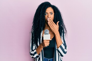 Middle age african american woman holding reporter microphone covering mouth with hand, shocked and...