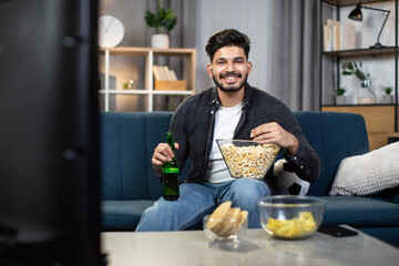 Positive indian guy in casual outfit enjoying tasty snacks and cold beer during football viewing on TV. Concept of people, leisure time and sport.