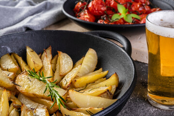 Roasted potatoes with rosemary in iron casserole and plate of confit tomatoes on dark background