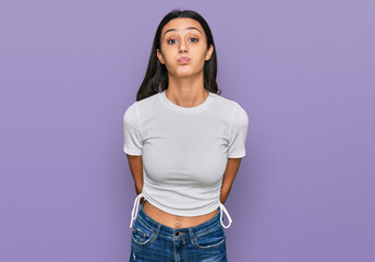 Young hispanic girl wearing casual white t shirt puffing cheeks with funny face. mouth inflated...