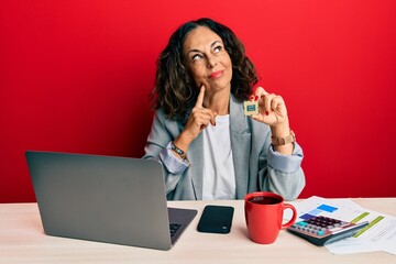 Beautiful middle age woman holding cpu computer processor serious face thinking about question with hand on chin, thoughtful about confusing idea