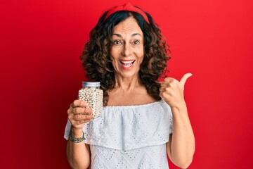 Middle age hispanic woman holding jar of beans pointing thumb up to the side smiling happy with open mouth