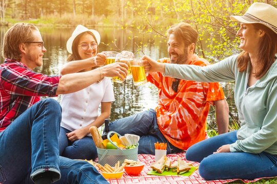 Happy group of friends drinking beers in the park during a pic nic barbecue - Couples of friends cheering together by the lake - Lifestyle and people concept.