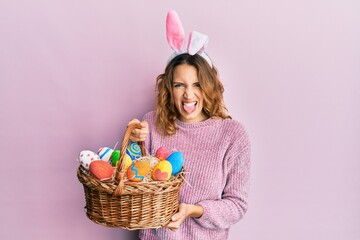Young caucasian woman wearing cute easter bunny ears holding colored egg sticking tongue out happy...
