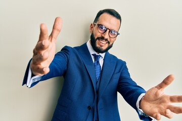 Young man with beard wearing business suit and tie looking at the camera smiling with open arms for hug. cheerful expression embracing happiness.