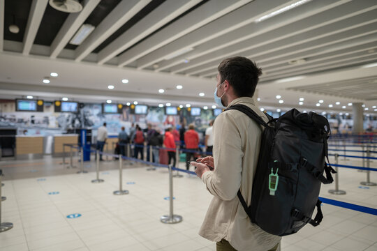 Man In Protective Mask With Phone At Airport Departure Check-in Desk For Baggage Drop-off At Terminal And Check-in For Flight, Maintains Social Distance. Travel During Covid 19 Pandemic. New Normal