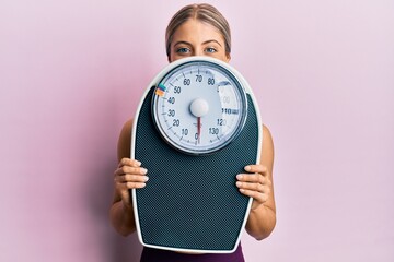 Beautiful blonde woman holding weight machine to balance weight loss smiling and laughing hard out...