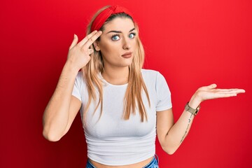 Young caucasian woman wearing casual white t shirt confused and annoyed with open palm showing copy space and pointing finger to forehead. think about it.