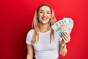 Young caucasian woman holding czech koruna banknotes looking positive and happy standing and smiling with a confident smile showing teeth