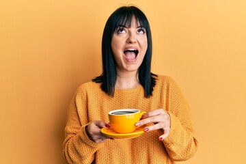 Young hispanic woman holding coffee angry and mad screaming frustrated and furious, shouting with...