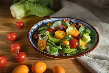 Vegetarian salad of pac choi cabbage, kiwi, tomatoes, kumquat, microgreen sprouts on a wooden background. Side view, close up, hard light.
