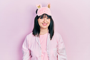 Young hispanic woman wearing sleep mask and pajama looking positive and happy standing and smiling with a confident smile showing teeth