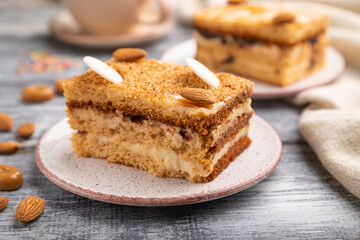 Honey cake with milk cream, caramel, almonds and a cup of coffee on a gray wooden background. Side view, selective focus.