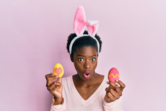 Young african american girl wearing cute easter bunny ears holding painted eggs afraid and shocked with surprise and amazed expression, fear and excited face.