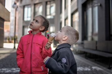 Fototapeta premium Pretty kids eating ice cream at street over urban background, funny candy girl enjoy sweet dessert and share it with her brother, street food, outside lifestyle