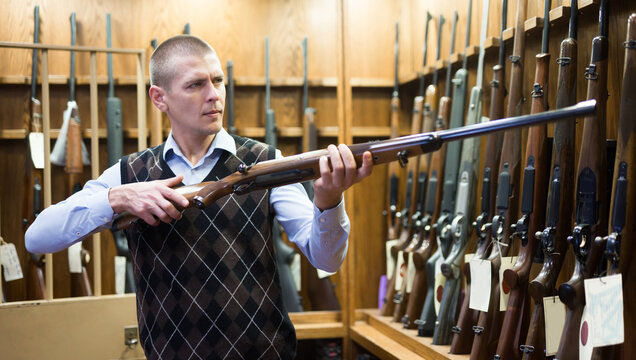 Confident male hunter examining hunting shotgun before buying in gun store..
