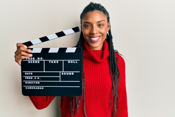 African american woman with braids holding video film clapboard looking positive and happy standing...