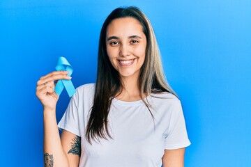 Young hispanic woman holding blue ribbon looking positive and happy standing and smiling with a confident smile showing teeth