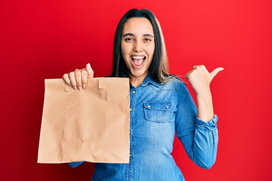 Young hispanic woman holding take away paper bag pointing thumb up to the side smiling happy with open mouth