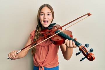 Beautiful brunette little girl playing violin celebrating crazy and amazed for success with open eyes screaming excited.