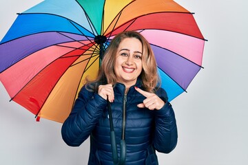 Middle age caucasian woman holding colorful umbrella smiling happy pointing with hand and finger