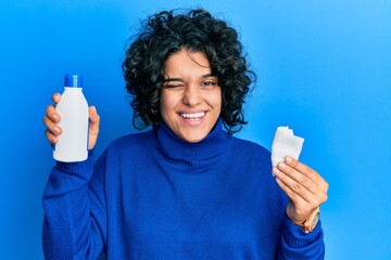 Young hispanic woman with curly hair holding medical alcohol winking looking at the camera with...