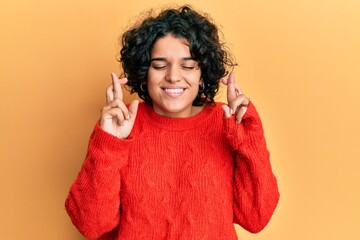Young hispanic woman with curly hair wearing casual winter sweater gesturing finger crossed smiling with hope and eyes closed. luck and superstitious concept.