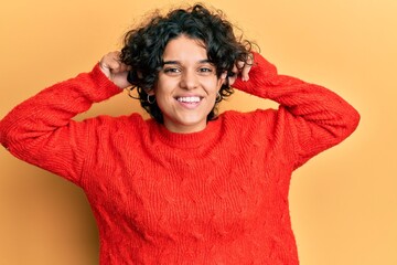 Young hispanic woman with curly hair wearing casual winter sweater smiling pulling ears with...