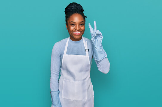 Young african american woman wearing apron smiling with happy face winking at the camera doing victory sign. number two.