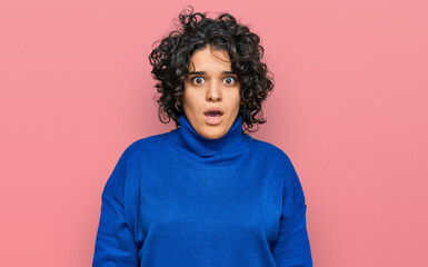 Young hispanic woman with curly hair wearing turtleneck sweater in shock face, looking skeptical and sarcastic, surprised with open mouth