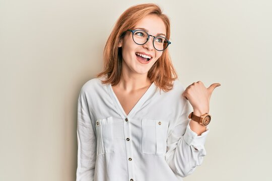 Young caucasian woman wearing casual clothes and glasses pointing thumb up to the side smiling happy with open mouth