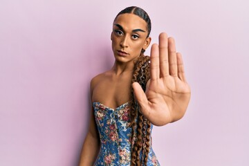 Hispanic man wearing make up and long hair wearing elegant corset doing stop sing with palm of the hand. warning expression with negative and serious gesture on the face.