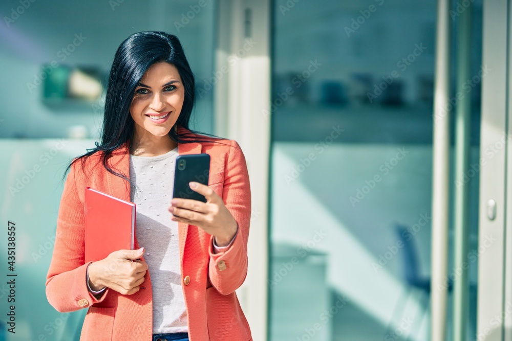 Wall mural Young beautiful businesswoman smiling happy using smartphone and holding book at the city.