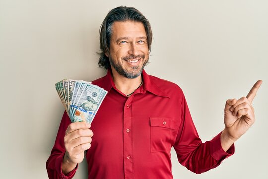 Middle age handsome man holding dollars smiling happy pointing with hand and finger to the side
