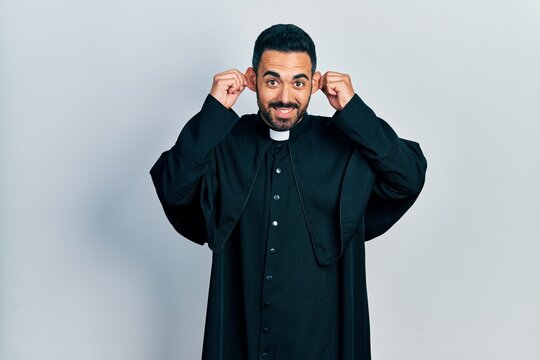 Handsome hispanic man with beard wearing catholic priest robe smiling pulling ears with fingers, funny gesture. audition problem