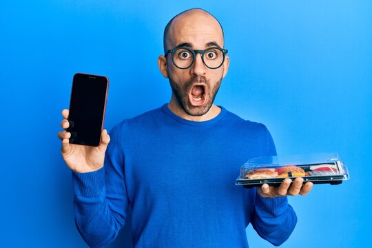 Young hispanic man holding take away food showing smartphone screen afraid and shocked with surprise and amazed expression, fear and excited face.