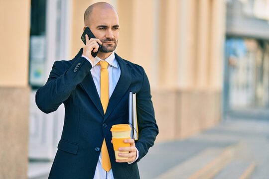 Young hispanic bald businessman with serious expression talking on the smartphone drinking coffee at the city