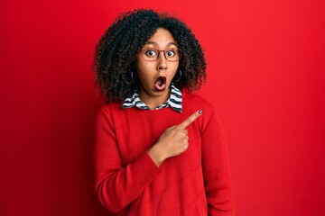 Beautiful african american woman with afro hair wearing sweater and glasses surprised pointing with...