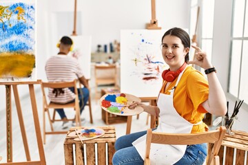 Young artist woman painting on canvas at art studio smiling and confident gesturing with hand doing small size sign with fingers looking and the camera. measure concept.