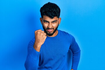 Arab man with beard wearing casual blue sweater angry and mad raising fist frustrated and furious while shouting with anger. rage and aggressive concept.