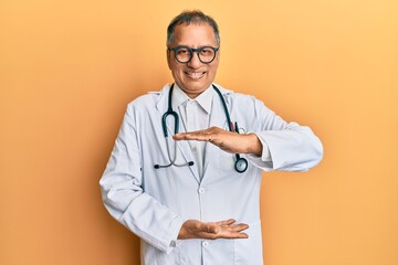 Middle age indian man wearing doctor coat and stethoscope gesturing with hands showing big and large size sign, measure symbol. smiling looking at the camera. measuring concept.