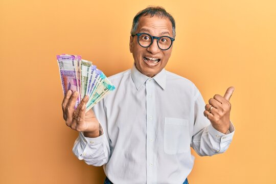 Middle age indian man holding indian rupee banknotes pointing thumb up to the side smiling happy with open mouth
