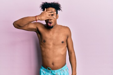 Fototapeta premium Young african american man with beard wearing swimsuit peeking in shock covering face and eyes with hand, looking through fingers with embarrassed expression.
