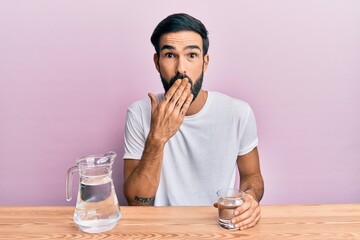 Young hispanic man drinking glass of water sitting on the table covering mouth with hand, shocked...