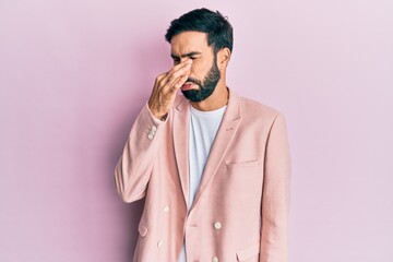 Young hispanic man wearing business jacket smelling something stinky and disgusting, intolerable smell, holding breath with fingers on nose. bad smell