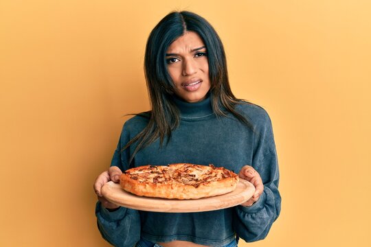 Young latin transsexual transgender woman holding italian pizza clueless and confused expression. doubt concept.